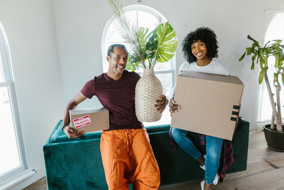 A young woman and a man are sitting on a wooden floor indoors, engaged in packing household items in preparation for a home relocation. The woman is holding a small bubble-wrapped item, while the man
